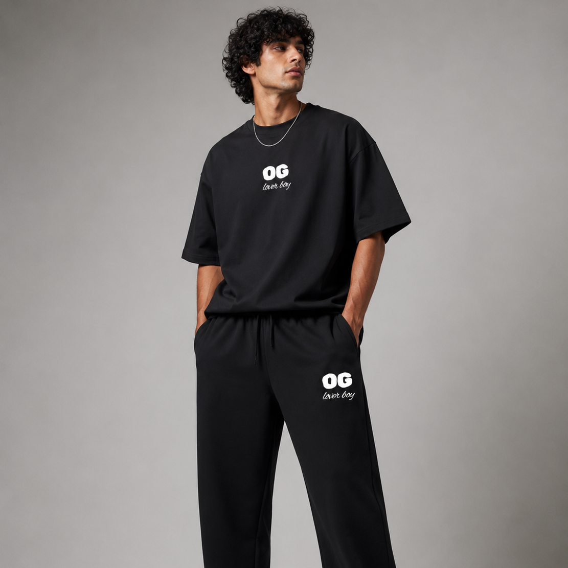 A man modeling a black oversized t-shirt and matching sweatpants set from the brand OG Day, featuring a white printed logo on the chest and thigh, paired with black sneakers, against a neutral gray studio background.